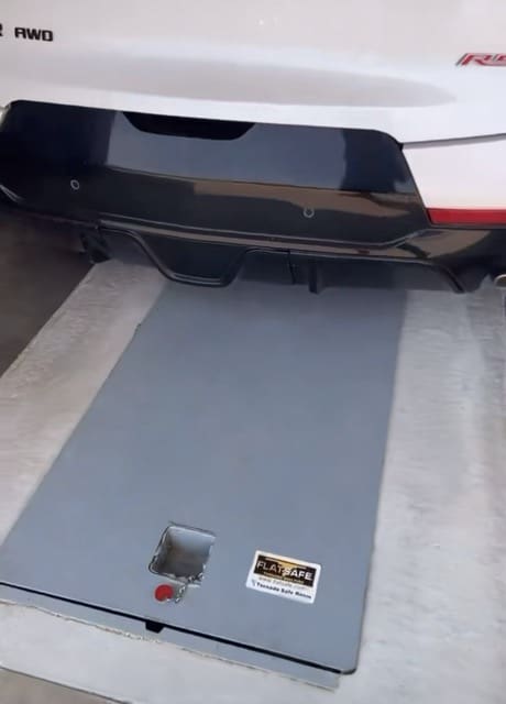 Vehicle parked over a closed galvanized underground garage storm shelter with a flush-mounted steel door installed in a residential garage.